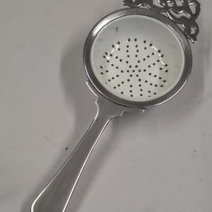 Può includere: Un colino da tè argentato con un manico decorativo e un filtro a forma di ciotola. Il filtro ha molti piccoli fori. Il manico ha un design curvo. Il colino è progettato per l'infusione di tè sfuso.