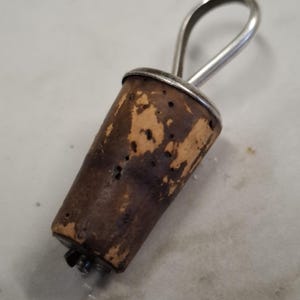 May include: A tapered brown cork bottle stopper featuring a natural porous texture and a silver-toned metal loop handle attached to the top. The stopper has a cylindrical shape that narrows towards the bottom, with a small metal component visible at the base.