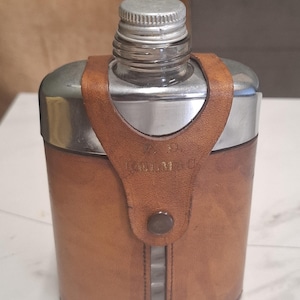 May include: A silver-toned metal flask encased in a tan leather sleeve with a vertical cutout revealing the glass body. The leather features a strap with a snap button closure and the embossed text P. D. KULMAO. It has a threaded silver metal cap.