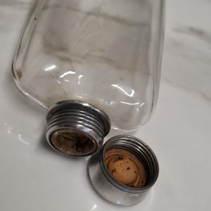 Pode incluir: Uma garrafa de vidro transparente com uma tampa de rosca de metal prateado e uma tampa separada a condizer com uma rolha de cortiça. A garrafa está deitada de lado, com a tampa desaparafusada e ao lado. A garrafa está vazia.