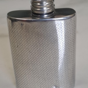 May include: A silver-toned metal flask with a curved, rectangular body and a textured, repeating geometric pattern across its surface. The flask features a cylindrical, screw-top cap with a ribbed edge and a smooth, polished finish at the neck.