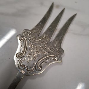 Könnte beinhalten: Eine silberne Gabel mit einem verzierten, gravierten Griff. Die Gabel hat drei Zinken und einen langen, schlanken Griff.