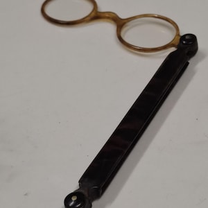 Op de afbeelding: Antieke opvouwbare bril met een schildpadmontuur en donkerbruin handvat. De ronde lenzen zijn gezet in een lichtbruin montuur. Het handvat kan worden opgevouwen voor compacte opslag. Een vintage accessoire.