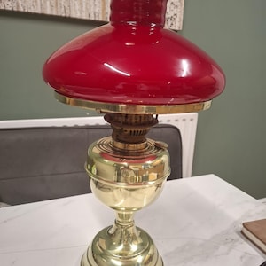 May include: A kerosene lamp featuring a polished brass base and a wide, rounded red glass shade. The lamp has a central brass burner mechanism connecting the base to the glass top, with a smooth, reflective metallic finish on the lower body.