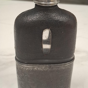 Puede incluir: Una petaca con una sección superior de cuero texturizado marrón oscuro y una ventana vertical plateada. La parte inferior es de metal plateado con un grabado floral intrincado. Cuenta con un tapón de rosca de color plateado.