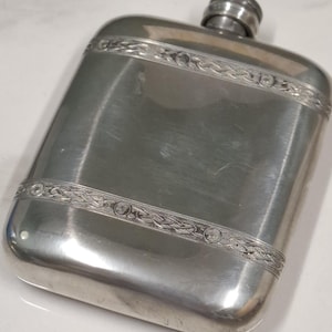 May include: A rectangular silver-toned metal flask with rounded corners and a screw-top cap. The flask features two horizontal bands of intricate, embossed Celtic-style knotwork patterns that span the width of the front surface.