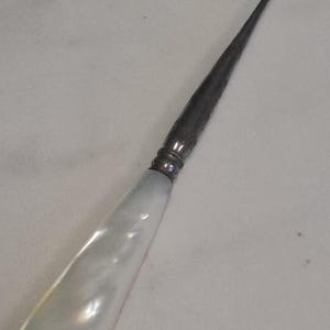 Op de afbeelding: Een haaknaald met een glad, iriserend wit parelmoeren handvat. Het handvat is verbonden met een lange, slanke, donker metalen schacht die taps toeloopt naar een kleine, gebogen haak aan de punt, ontworpen voor gedetailleerd handwerk.