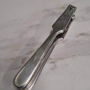 Peut inclure: Un casse-noix en métal argenté doté d'une conception articulée avec deux longs manches lisses et une zone de préhension texturée à motif en losange près de la charnière supérieure. L'outil présente une finition polie et une forme effilée.