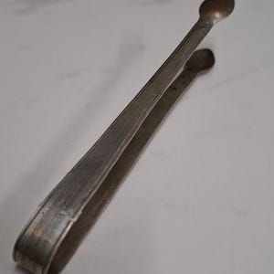 Könnte beinhalten: Silberne Zuckerzange mit abgerundetem, löffelartigem Ende. Die Zange ist aus Metall und hat eine strukturierte Oberfläche.