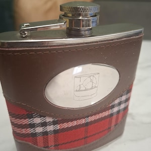May include: A metal flask featuring a brown leather-effect wrap with a red, black, and white plaid fabric insert. The front has an oval silver-toned plate engraved with a line drawing of a glass containing ice cubes and a screw-top metal cap.