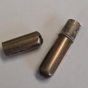 Puede incluir: Dos objetos cilíndricos antiguos de latón, uno abierto y otro cerrado. Los objetos tienen un aspecto deslustrado, lo que sugiere antigüedad. El cilindro cerrado tiene un extremo redondeado y una banda texturizada cerca de la parte superior. El cilindro abierto tiene un extremo cortado.