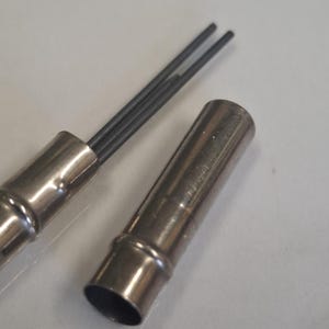 Op de afbeelding: Een zilveren mechanische potloodhouder met drie donkergrijze vullingen. De dop is losgemaakt, waardoor de vullingen zichtbaar zijn. De potloodhouder is gemaakt van glanzend metaal.