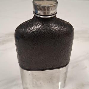 Könnte beinhalten: Ein Taschenflachmann mit einem dunkelbraunen, strukturierten Lederüberzug im oberen Bereich und einem polierten, silberfarbenen Metallsockel. Der Flachmann hat einen passenden silberfarbenen Schraubverschluss mit geriffeltem Rand am Flaschenhals.