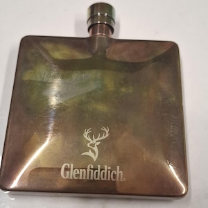 May include: A square-shaped metal flask with a polished bronze finish. The front surface features an engraved stag logo and the text Glenfiddich. The flask includes a cylindrical screw-top cap at the centre of the top edge, reflecting light.
