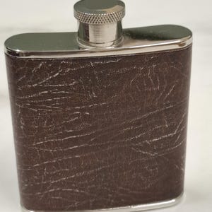 Puede incluir: Una petaca rectangular de metal con un revestimiento marrón oscuro texturizado con un patrón de grano visible. La petaca tiene un marco de metal plateado pulido en la parte superior e inferior, y una tapa de rosca plateada con borde estriado.