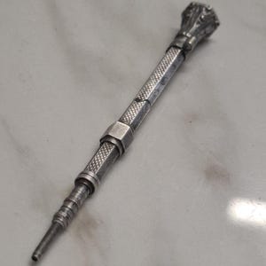 Op de afbeelding: Een zilverkleurig metalen gereedschap met een getextureerde, cilindrische handgreep met ruitpatroon, een gladde zeshoekige kraag en een taps toelopende, geribbelde punt. Het boveneinde is afgewerkt met een decoratieve, veelzijdige knop met details.