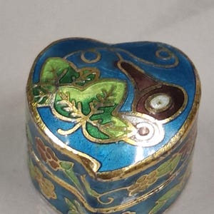 May include: Heart-shaped trinket box with a blue enamel finish, accented with gold trim and floral designs. The lid features green leaves and a brown, circular design. A decorative piece.
