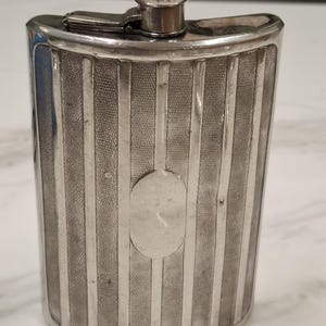 May include: A silver-toned metal flask with a curved, rectangular shape. The front surface features a repeating pattern of vertical polished stripes alternating with textured, cross-hatched panels. A smooth, blank oval plaque is centred on the front. The top includes a hinged, screw-down cap.