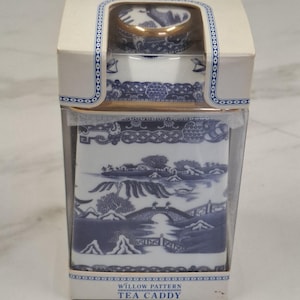 Könnte beinhalten: Eine quadratische Teedose mit blau-weißem Willow-Muster, verpackt in einem cremefarbenen Karton mit transparentem Kunststofffenster. Der Karton trägt oben und unten den Schriftzug Willow Pattern Tea Caddy, wobei ein goldfarbener Deckel sichtbar ist.