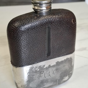 Puede incluir: Una petaca rectangular con una sección superior de cuero texturizado marrón oscuro que incluye una ventana de visualización vertical de vidrio y una sección inferior de metal plateado pulido con un monograma grabado y tapa de rosca.