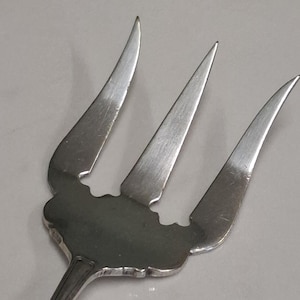 Puede incluir: Un tenedor de servir plateado con tres púas. El tenedor tiene un borde decorativo y un mango largo y delgado. Las púas son afiladas y curvadas. El utensilio probablemente está hecho de acero inoxidable.
