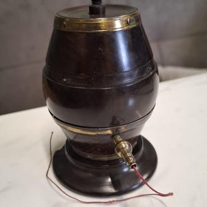 Puede incluir: Un recipiente en forma de barril, marrón oscuro, con detalles de latón y un pequeño pomo de madera. Tiene un grifo y un cordón rojo. El recipiente está sobre una superficie blanca. Objeto vintage.