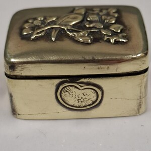 May include: A small, rectangular brass box with a hinged lid. The lid features a detailed relief of a bird perched on a flowering branch. The front of the box has an oval design. The box has a vintage appearance.
