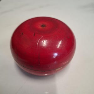 Op de afbeelding: Een rode houten bal met een gladde, glanzende afwerking. De bal heeft een kleine deuk aan de bovenkant.