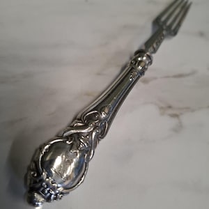 Op de afbeelding: Een zilveren vork met een sierlijke handgreep. De handgreep heeft gedetailleerd krulwerk en een afgerond uiteinde. De tanden van de vork zijn bovenaan zichtbaar. De vork is waarschijnlijk gemaakt van sterling zilver of een vergelijkbaar metaal.