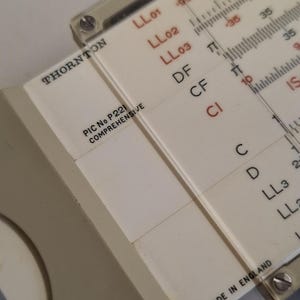 May include: Close-up of a vintage Thornton slide rule. The slide rule is off-white with black and red markings. The text "THORNTON" and "PIC No P221 COMPREHENSIVE" are visible. The slide rule is marked "MADE IN ENGLAND".