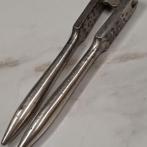 May include: A silver-toned metal nutcracker featuring two elongated, tapered handles and a hinged mechanism at the top. The tool has a textured grip area on the inner section of the handles and shows signs of wear with small dark spots on the surface.