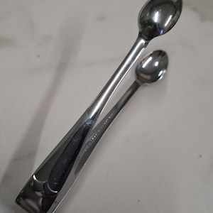 Könnte beinhalten: Eine silberfarbene Zuckerzange aus Metall mit zwei kleinen, ovalen Löffelenden, die durch einen flexiblen, gebogenen Griff verbunden sind. Die polierte Oberfläche reflektiert das Licht und unterstreicht das glatte, geschwungene Design dieses Küchenutensils.
