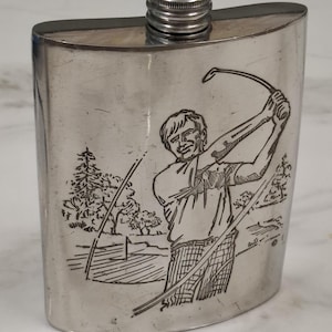 Op de afbeelding: Een zilverkleurige metalen heupfles met een gravure van een golfer tijdens een swing op een golfbaan. De fles heeft een gebogen rechthoekig lichaam met een schroefdop. De gedetailleerde ets toont een persoon in een shirt en een broek met patroon.