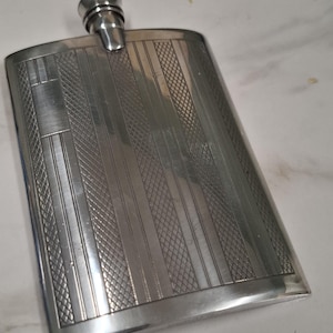 May include: A rectangular silver-coloured metal flask featuring a textured surface with alternating vertical stripes and cross-hatched patterns. The flask has a rounded top edge and a cylindrical screw-top cap centred at the top of the body.