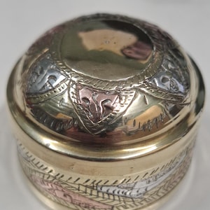 May include: A small, round brass trinket box with detailed etched patterns. The lid and base are gold, with silver and rose gold accents. The box features a decorative design around the sides and top, enhancing its ornate look. The box is likely used for storing small items.