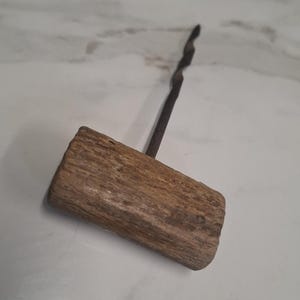 May include: A hand tool featuring a T-shaped handle made from a textured, light brown piece of wood attached to a long, thin, dark metal shaft with a twisted, spiral-like design extending from the centre of the wooden handle.