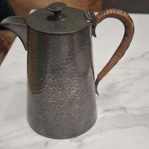 Könnte beinhalten: Ein hoher, zylindrischer Metallkrug mit gehämmertem Muster und dunkelgrauer Oberfläche. Er verfügt über einen Klappdeckel mit einem kleinen Knaufgriff und einen geschwungenen Seitengriff, der mit hellbraunem, geflochtenem Material umwickelt ist.