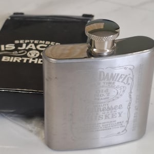 Pode incluir: Uma petaca de metal prateado com o logótipo gravado da Jack Daniel's Tennessee Whiskey na frente, acompanhada por uma bolsa preta com o texto SEPTEMBER IS JACK'S BIRTHDAY e o logótipo da marca Old No. 7 em letras brancas.