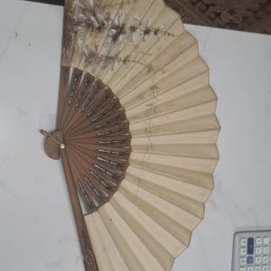 May include: A folding hand fan with a beige fabric leaf featuring a painted floral design in brown and white tones. The fan has a wooden frame with intricate cut-out details and a metal ring at the pivot point, showcasing a classic pleated structure.