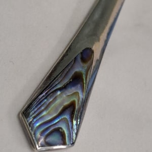 May include: Close-up of a silver-toned metal object with an abalone inlay. The inlay features iridescent blues, greens, and browns. The object has a geometric shape with a pointed end.