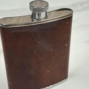 Puede incluir: Una petaca rectangular de metal con cuerpo curvo envuelto en cuero marrón oscuro. La petaca presenta una parte superior de metal plateado pulido, una base plateada a juego y un tapón de rosca circular con un borde texturizado y estriado.