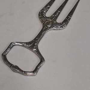 May include: Silver-toned serving fork with three tines and an ornate handle. The handle features detailed floral and scrollwork designs. The serving utensil is likely for cakes, pastries, or other desserts.