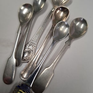 May include: Five silver teaspoons, one with a shell-shaped bowl, and one with an ornate handle. The spoons are arranged on a gray surface.