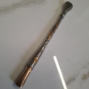May include: A silver-toned metal bombilla straw featuring a long, slender stem with intricate engraved floral patterns and gold-coloured accents. The straw has a smooth, plain copper-toned mouthpiece at one end and a flattened, flared filter tip at the other.
