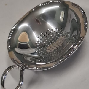 Puede incluir: Un colador de té ovalado de acero inoxidable brillante, con un asa. El colador tiene un cuenco perforado y un borde decorativo. El asa es un lazo simple y redondeado. La superficie refleja la luz, resaltando el metal pulido.