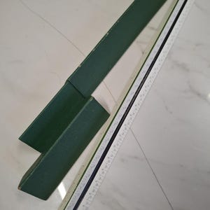 May include: A white and black architectural scale ruler with measurements, next to a dark green case. The ruler is marked with various units of measurement. The case is open, revealing the ruler.
