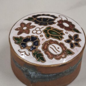 May include: A small, round trinket box. The white lid is decorated with a floral design in brown, blue, and green. The box has a bronze-coloured base with a dark blue band. The lid has gold accents. The box is approximately 5 cm in diameter.