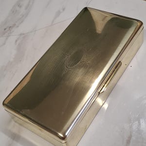 Könnte beinhalten: Eine rechteckige, goldfarbene Metallbox mit Scharnierdeckel. Die Box hat eine gebürstete Oberfläche und ein dekoratives ovales Design auf der Oberseite. Die Box wird wahrscheinlich zur Aufbewahrung kleiner Gegenstände verwendet.