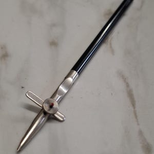 Puede incluir: Una herramienta de compás de dibujo con un mango negro largo y delgado unido a una punta de plata metálica. El mecanismo incluye una perilla de ajuste circular central con un pequeño acento rojo y un componente estabilizador de metal en forma de cruz.