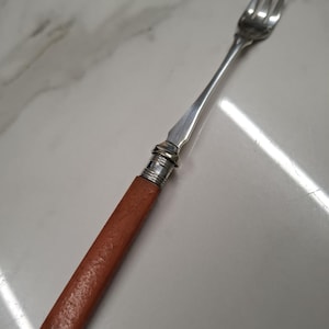 Op de afbeelding: Een zilveren serveervork met drie tanden en een bruin handvat. De handgreep heeft een getextureerde uitstraling en is met een zilveren band aan de vork bevestigd. De vork is ontworpen om te serveren.